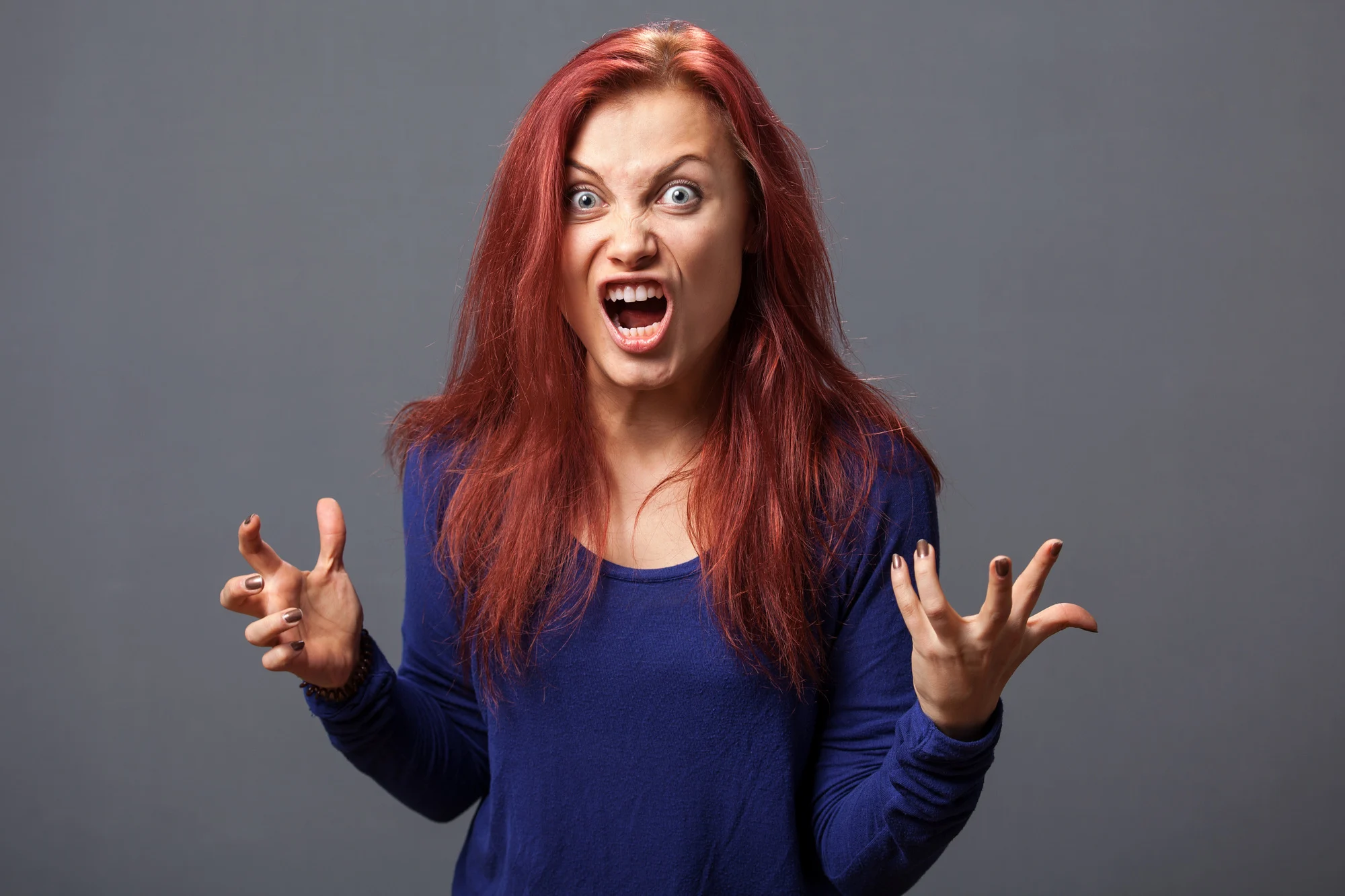 Young furious woman with opened mouth and mad look gesturing against gray background