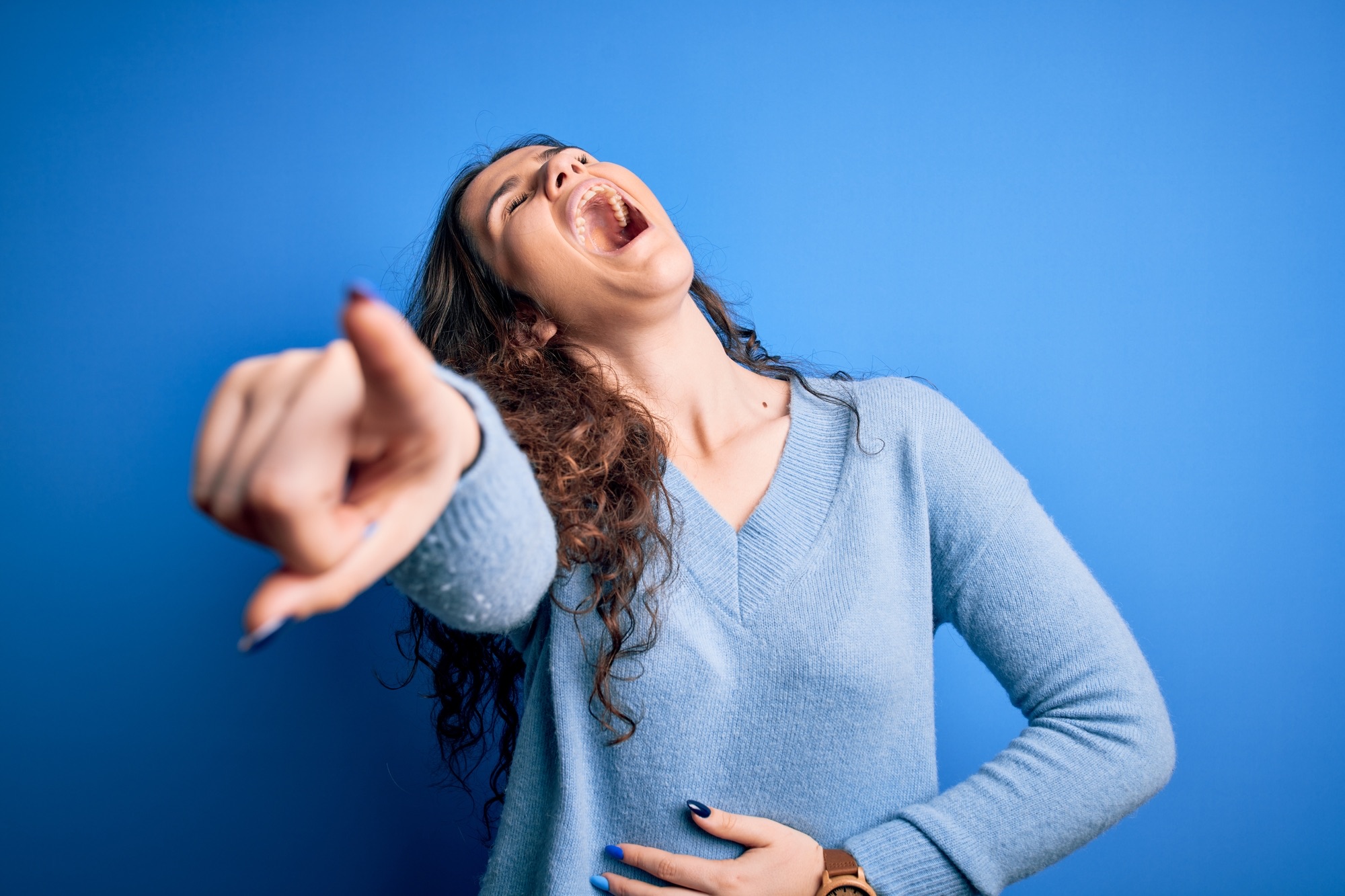 Young beautiful woman with curly hair wearing blue casual sweater over isolated background laughing at you, pointing finger to the camera with hand over body, shame expression