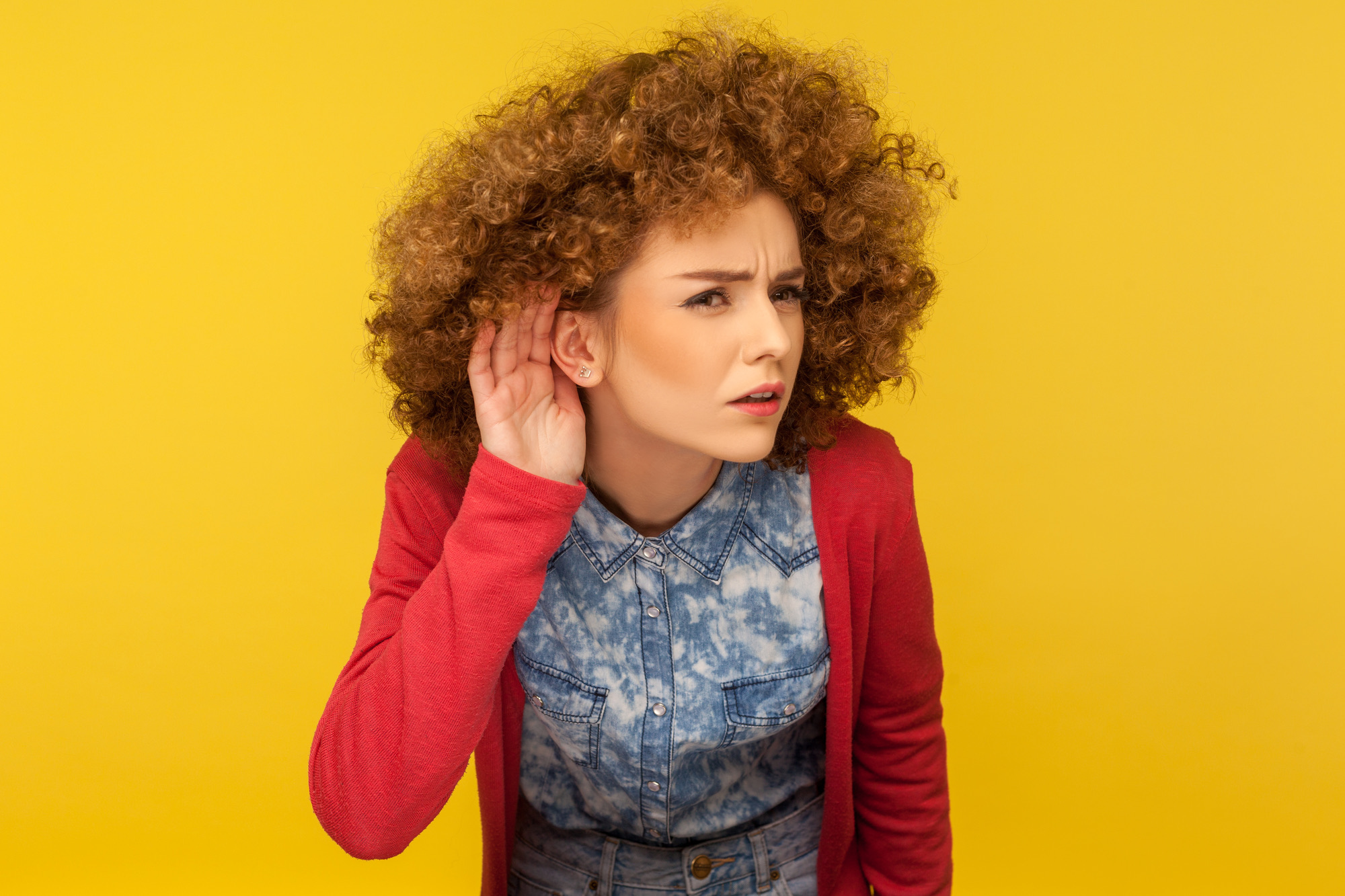 What are you saying? Portrait of woman with curly hair holding hand near ear and listening carefully intently, curious to hear secret quiet talk. indoor studio shot isolated on yellow background