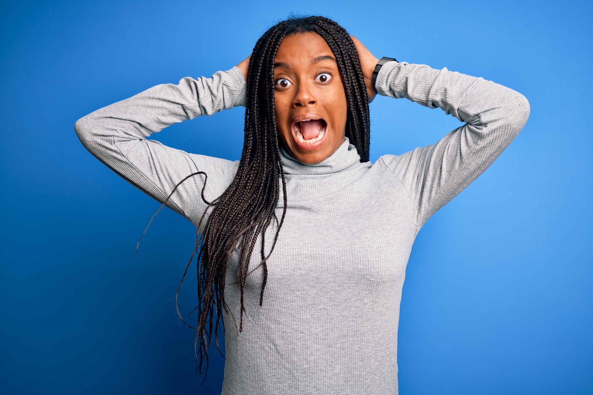 Young african american woman standing wearing casual turtleneck over blue isolated background Crazy and scared with hands on head, afraid and surprised of shock with open mouth