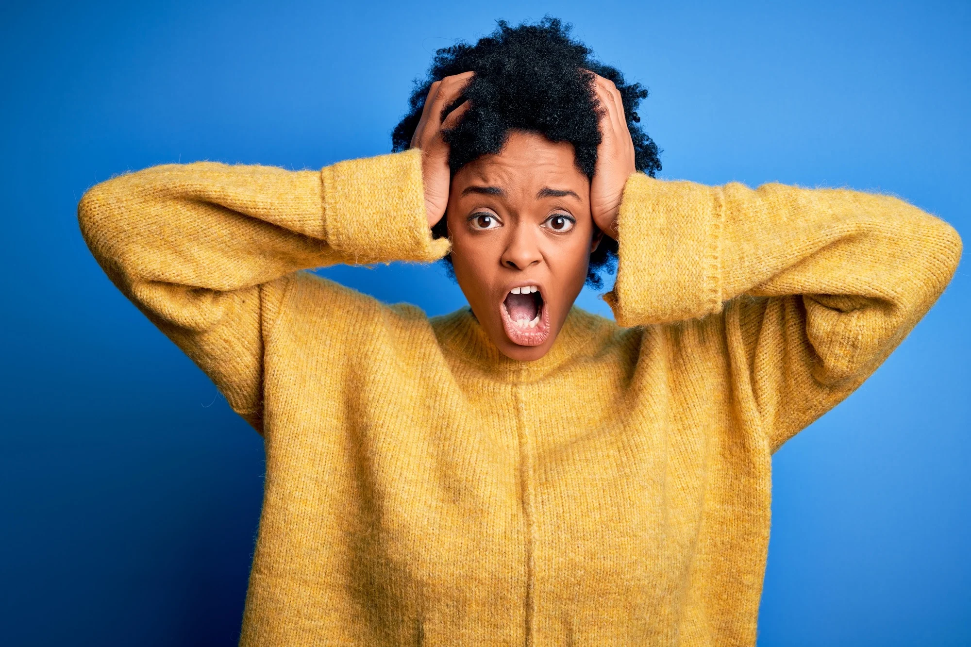 Young beautiful African American afro woman with curly hair wearing yellow casual sweater Crazy and scared with hands on head, afraid and surprised of shock with open mouth