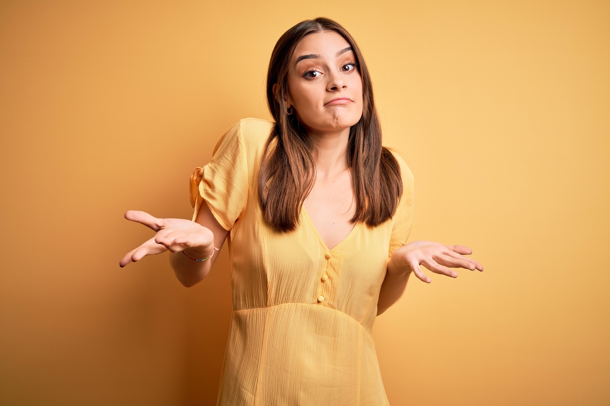 Young beautiful brunette woman wearing casual dress standing over yellow background clueless and confused expression with arms and hands raised. Doubt concept.