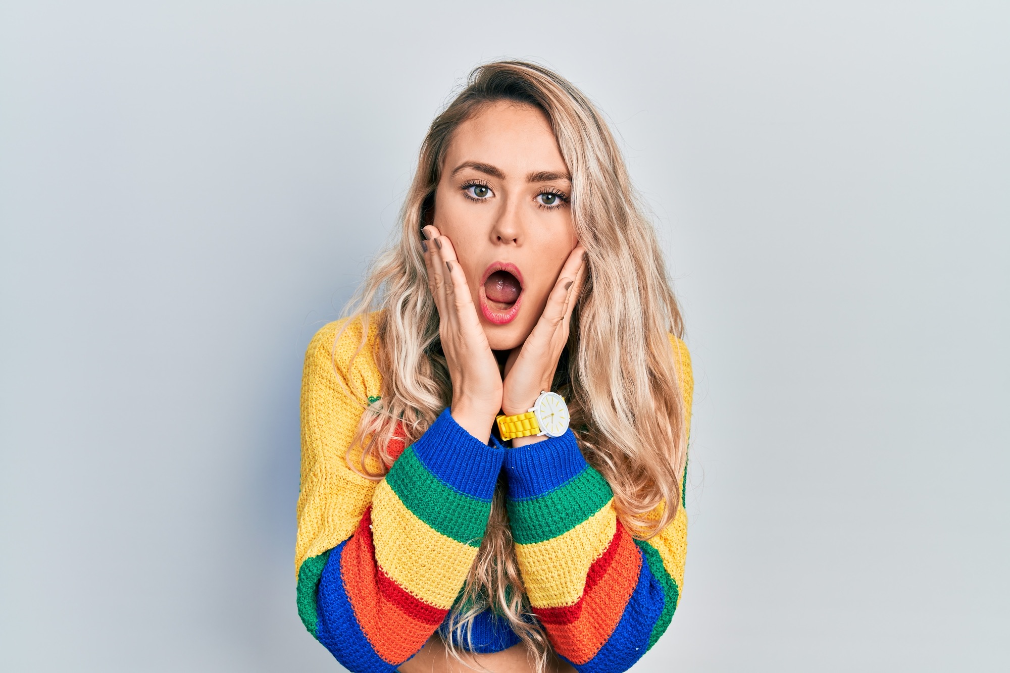 Beautiful young blonde woman wearing colored sweater afraid and shocked, surprise and amazed expression with hands on face