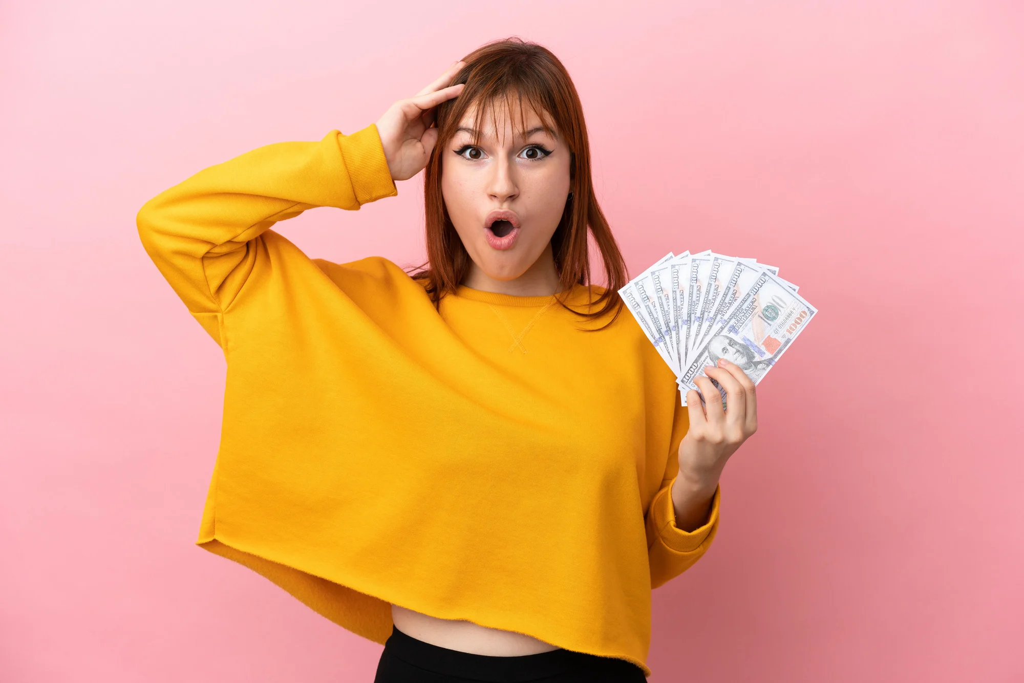 Redhead girl taking a lot of money isolated on pink background with surprise expression