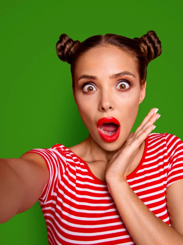 OOPS Self portrait of cute, trendy and shocked woman with bun hairdo wide open eyes mouth shooting selfie on front camera isolated on red background.