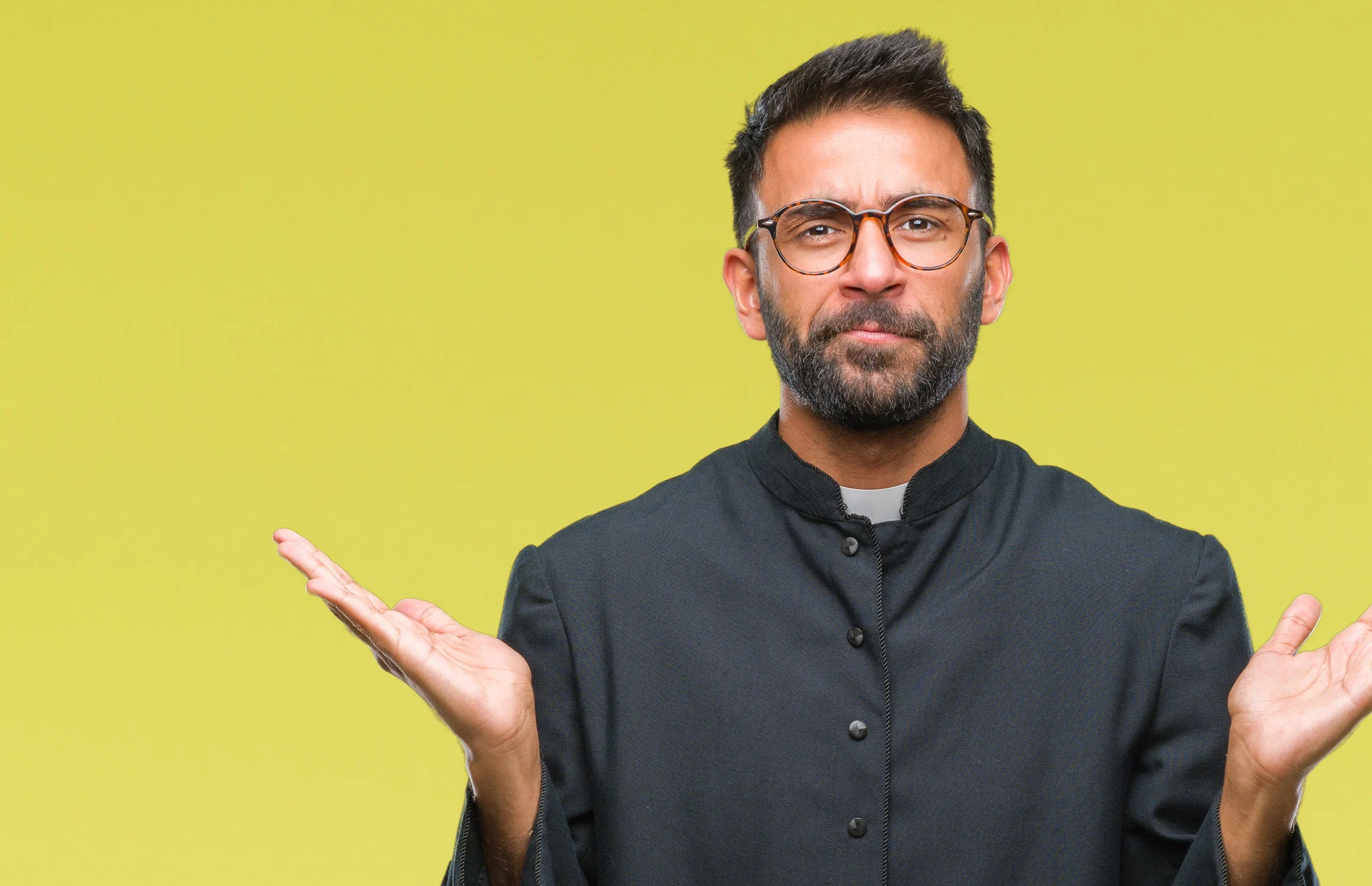 Adult hispanic catholic priest man over isolated background clueless and confused expression with arms and hands raised. Doubt concept.
