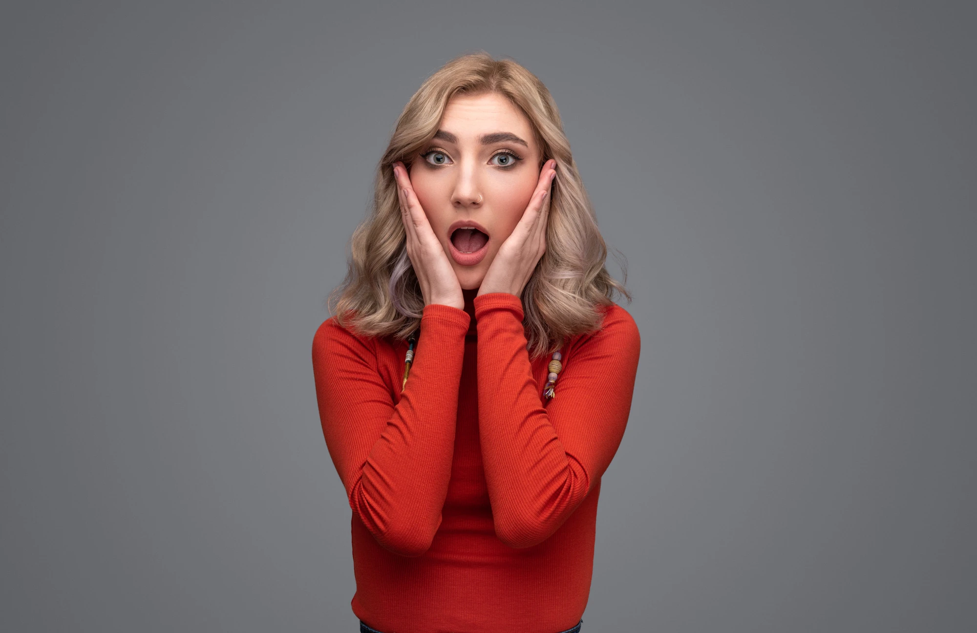 Pretty female in red turtleneck sweater keeping hands near cheeks and looking at camera with astonished face expression while standing against gray background