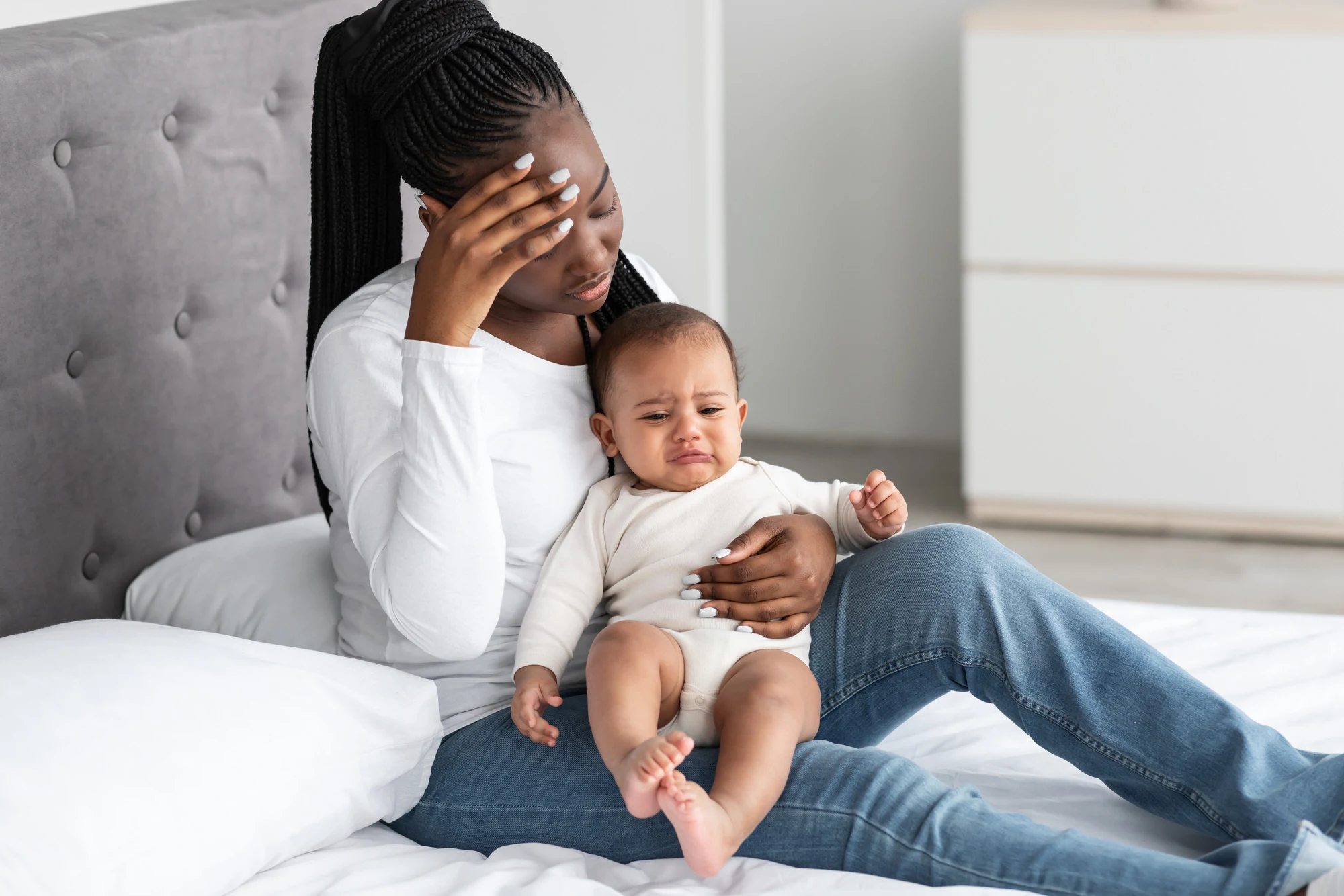 Postpartum Depression Concept. Portrait of tired African American woman sitting with her small black kid on bed at home, feeling headache, touching forehead with hand. Exhaustion And Parenthood