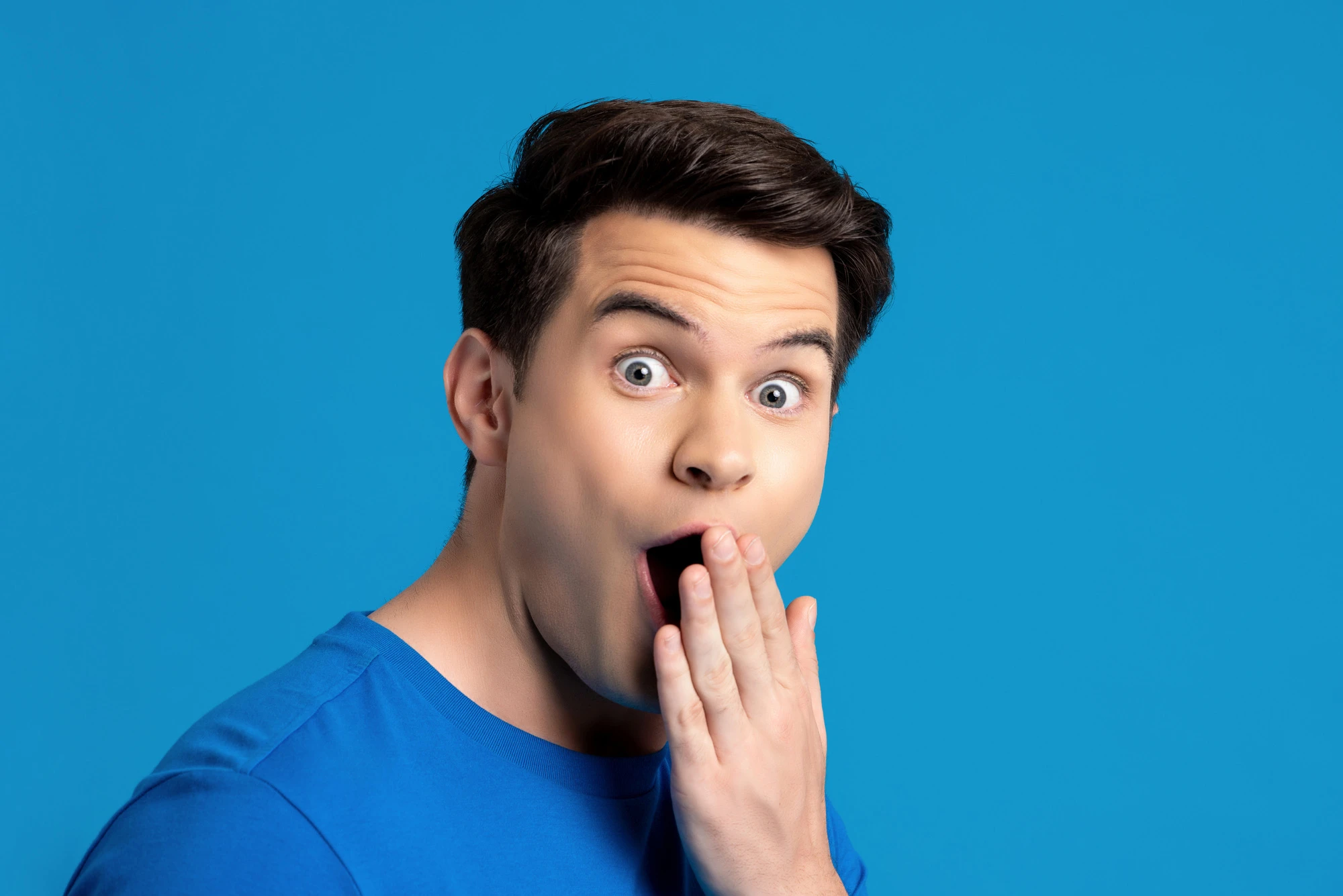 Portrait of caucasian male model in astonished gesture with hand covering mouth in blue isolated studio background, selective focused