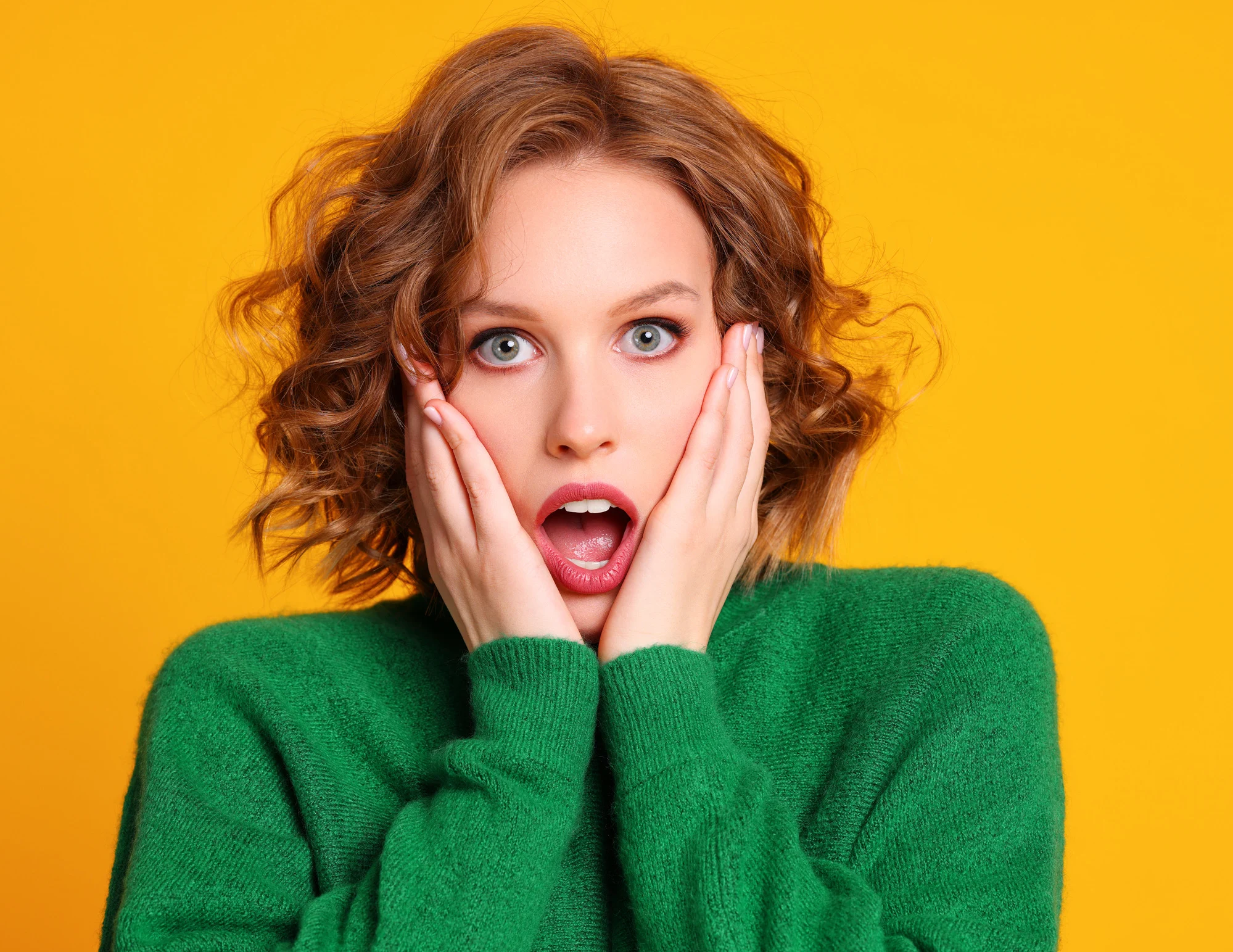 Amazed young woman in trendy outfit holding hands on cheeks and looking at camera against yellow background