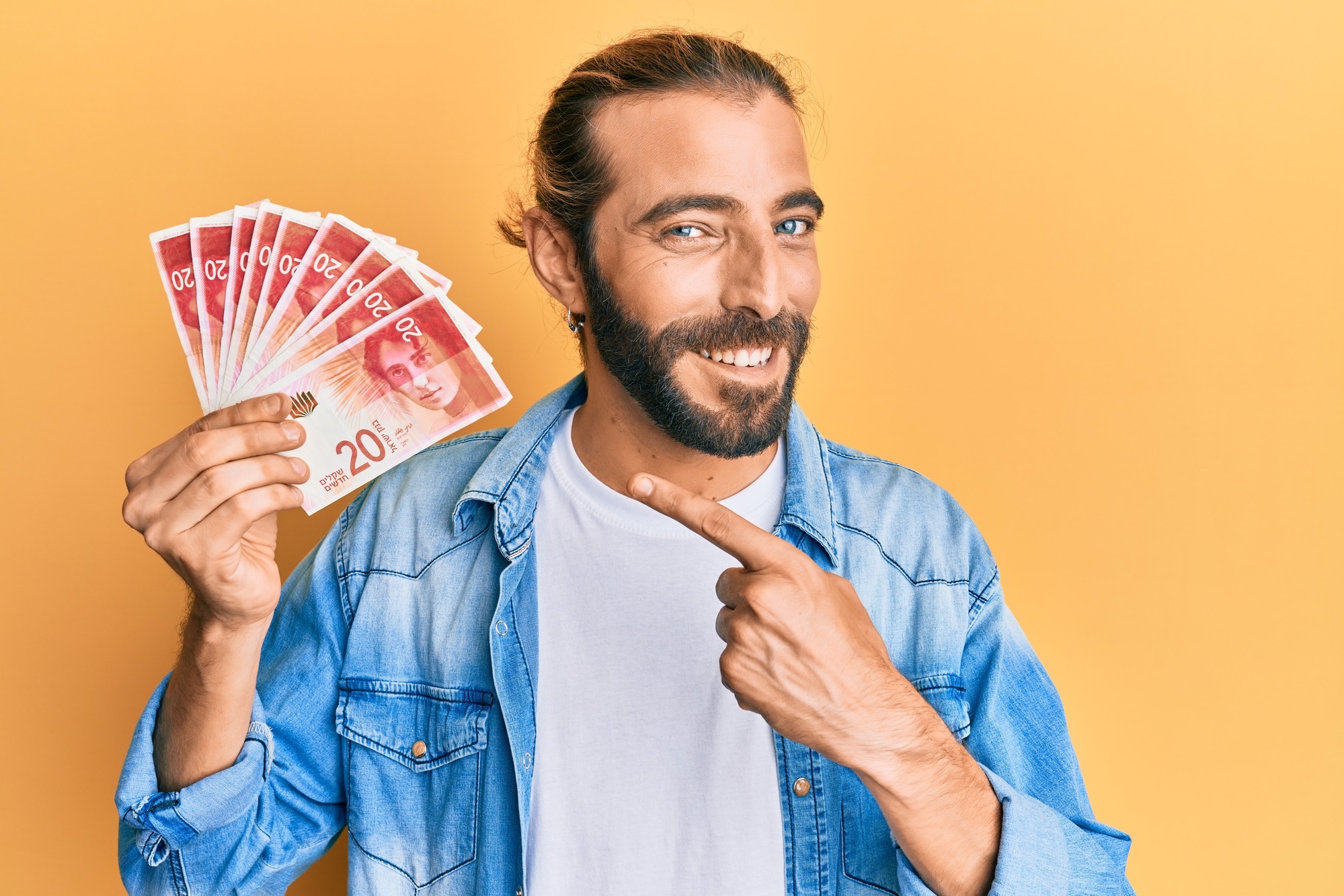 Attractive man with long hair and beard holding 20 israel shekels banknotes smiling happy pointing with hand and finger