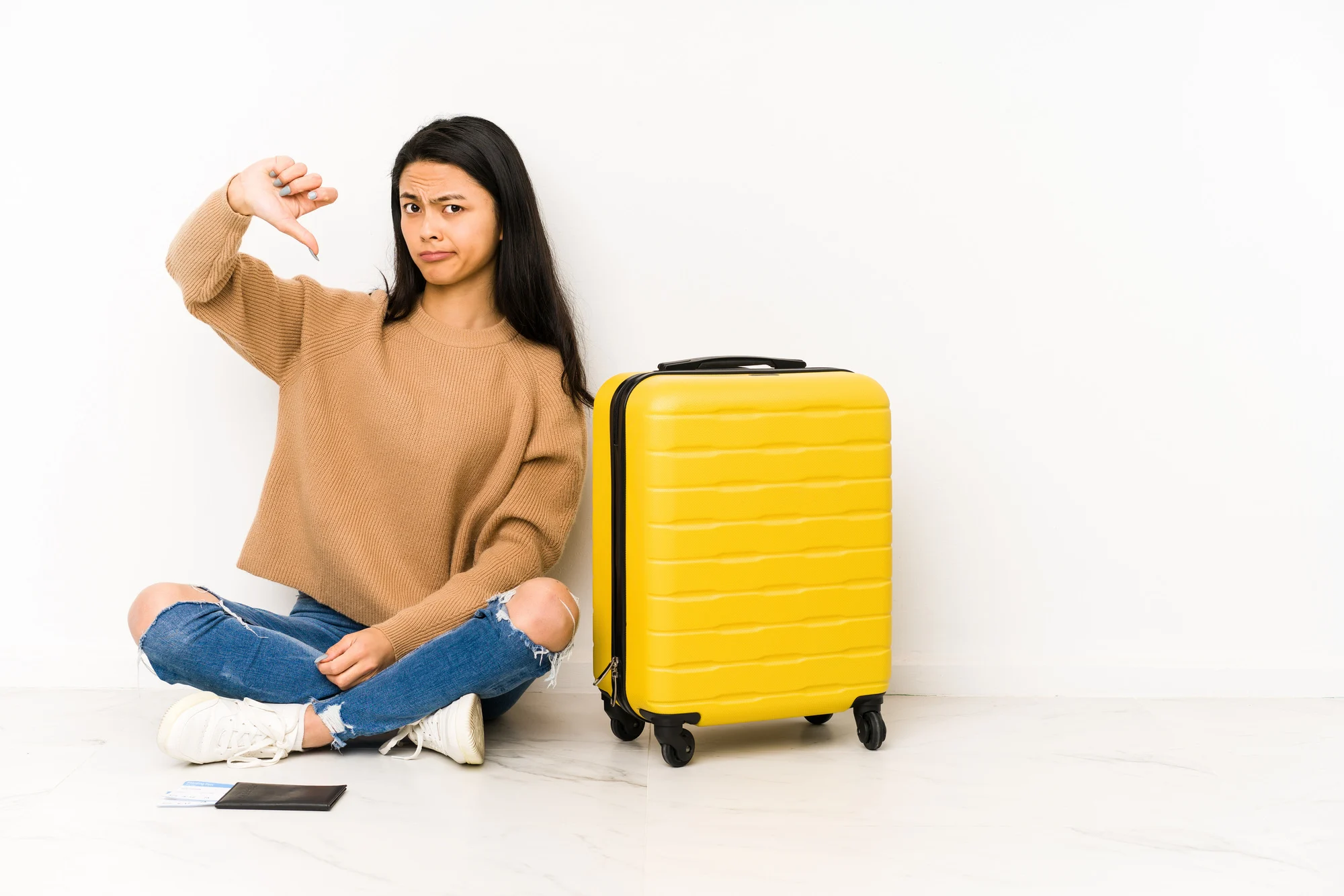 Young chinese traveler woman sittting on the floor with a suitcase isolated showing a dislike gesture, thumbs down. Disagreement concept.