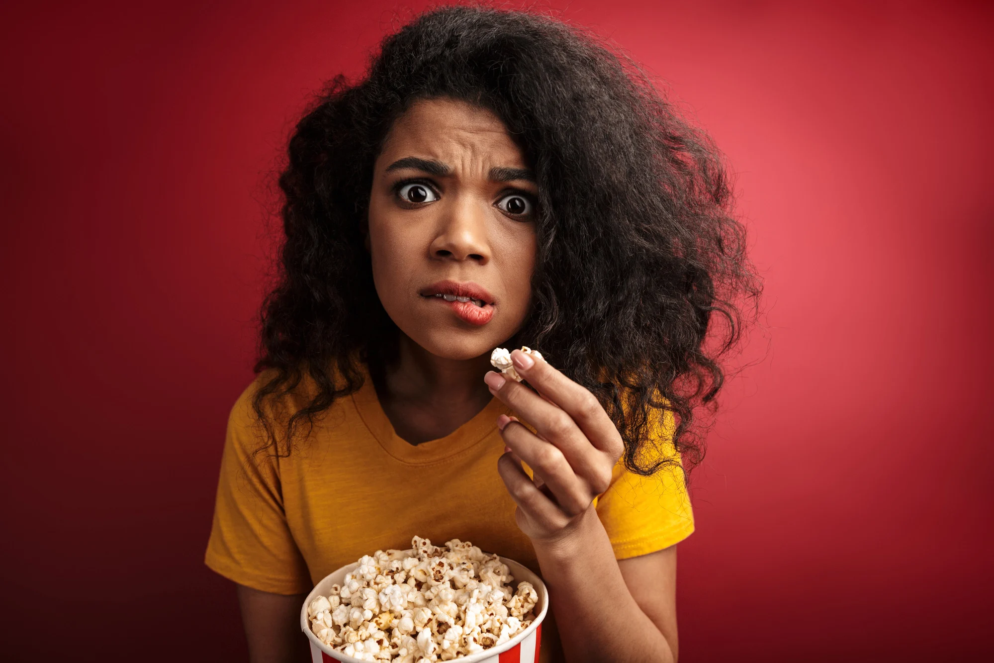 Image of shocked brunette african american woman with curly hair expressing wonder while holding popcorn bucket isolated over red background