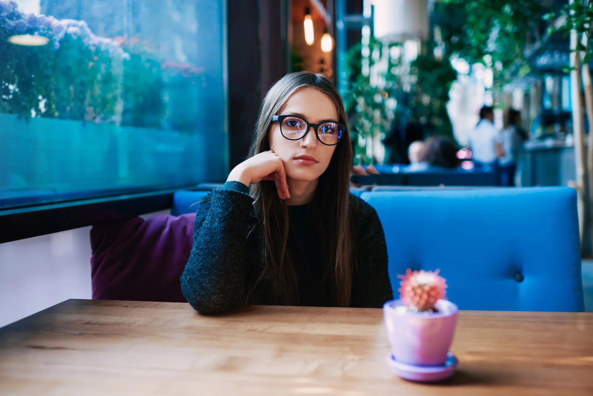 Wistfl female in casual clothes with eyeglasses looking at camera while sitting at wooden table with cactus in flower pot leaning on hand during rest
