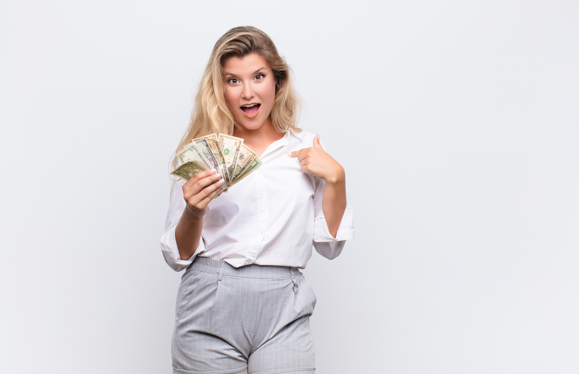 young pretty latin woman looking shocked and surprised with mouth wide open, pointing to self with dollar bills banknotes