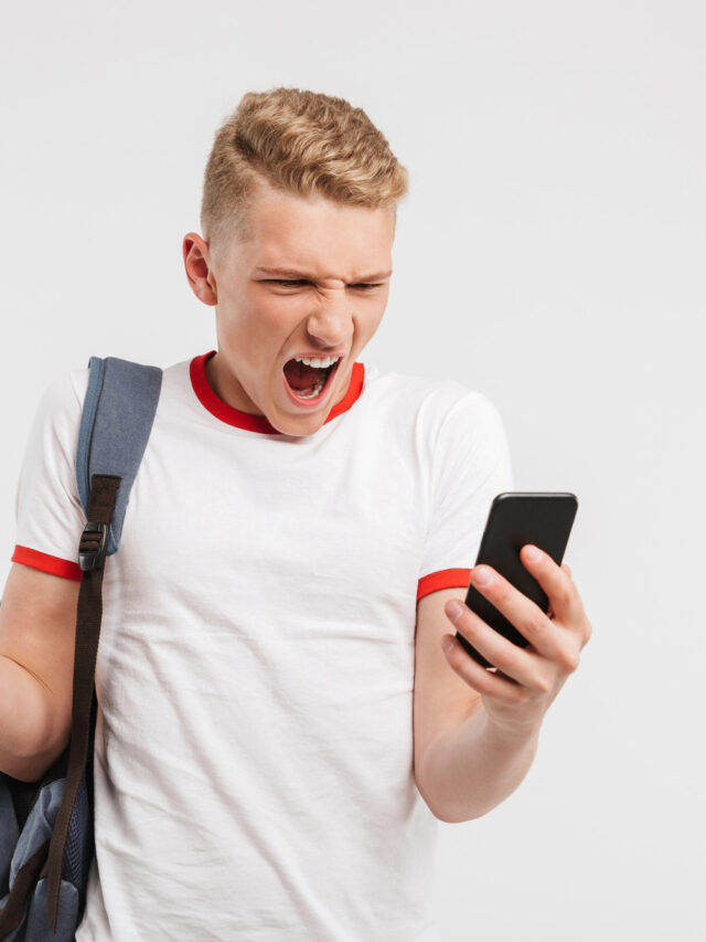 Portrait of an angry teenage boy standing with backpack and yelling at mobile phone isolated over white background