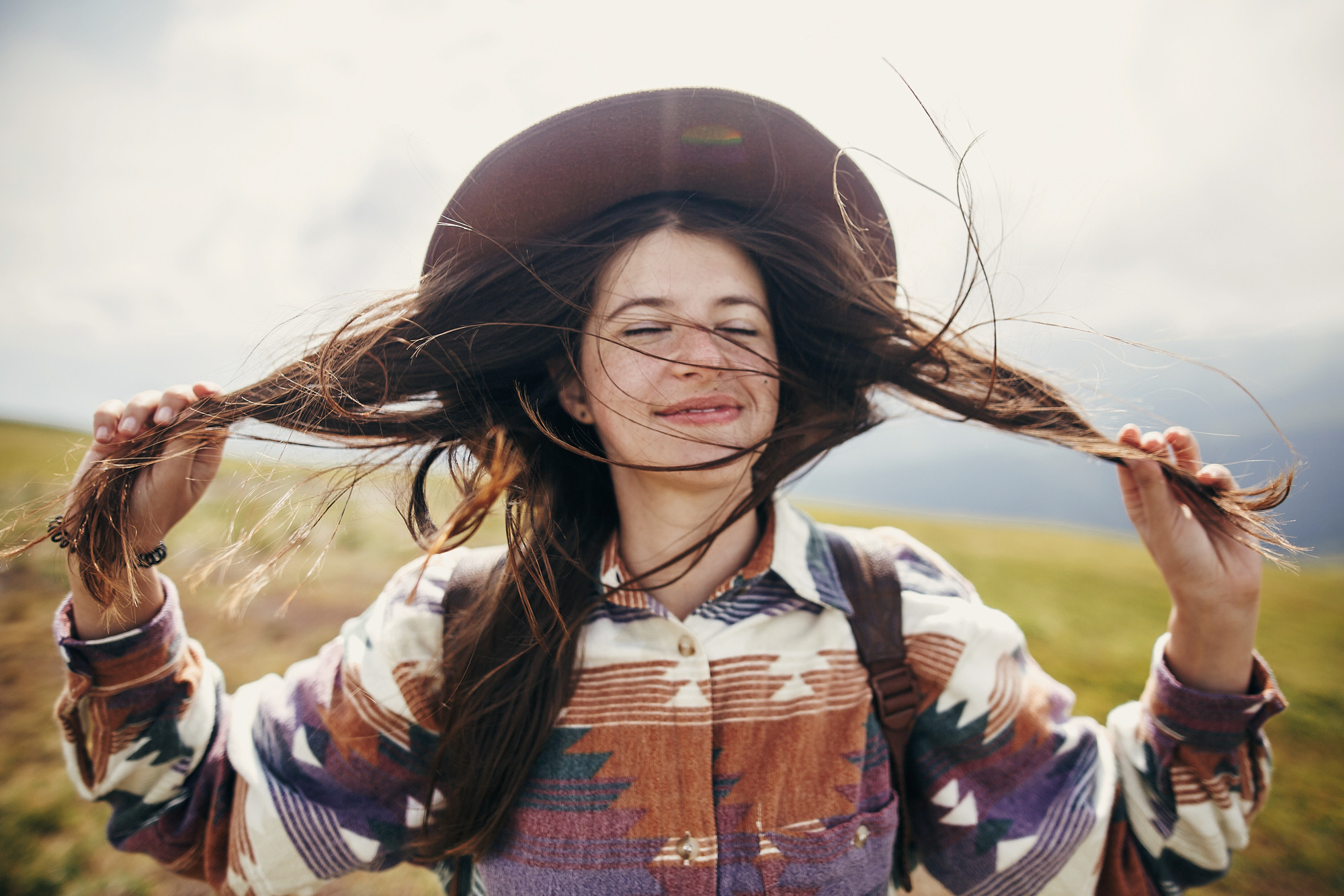 happy traveler hipster girl with windy hair smiling, standing on top of sunny mountains. space for text. stylish woman in hat holding hair. atmospheric moment. travel and wanderlust