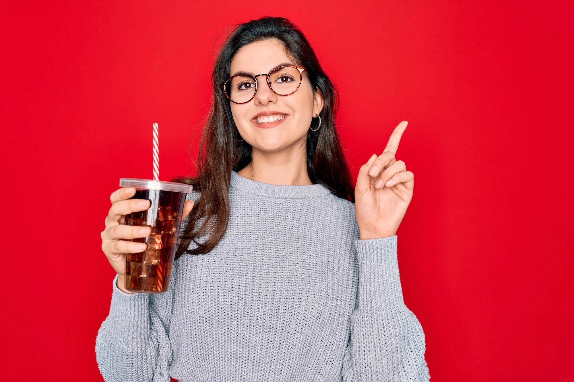 Young beautiful girl wearing glasses drinking sweet drink soda over red background surprised with an idea or question pointing finger with happy face, number one