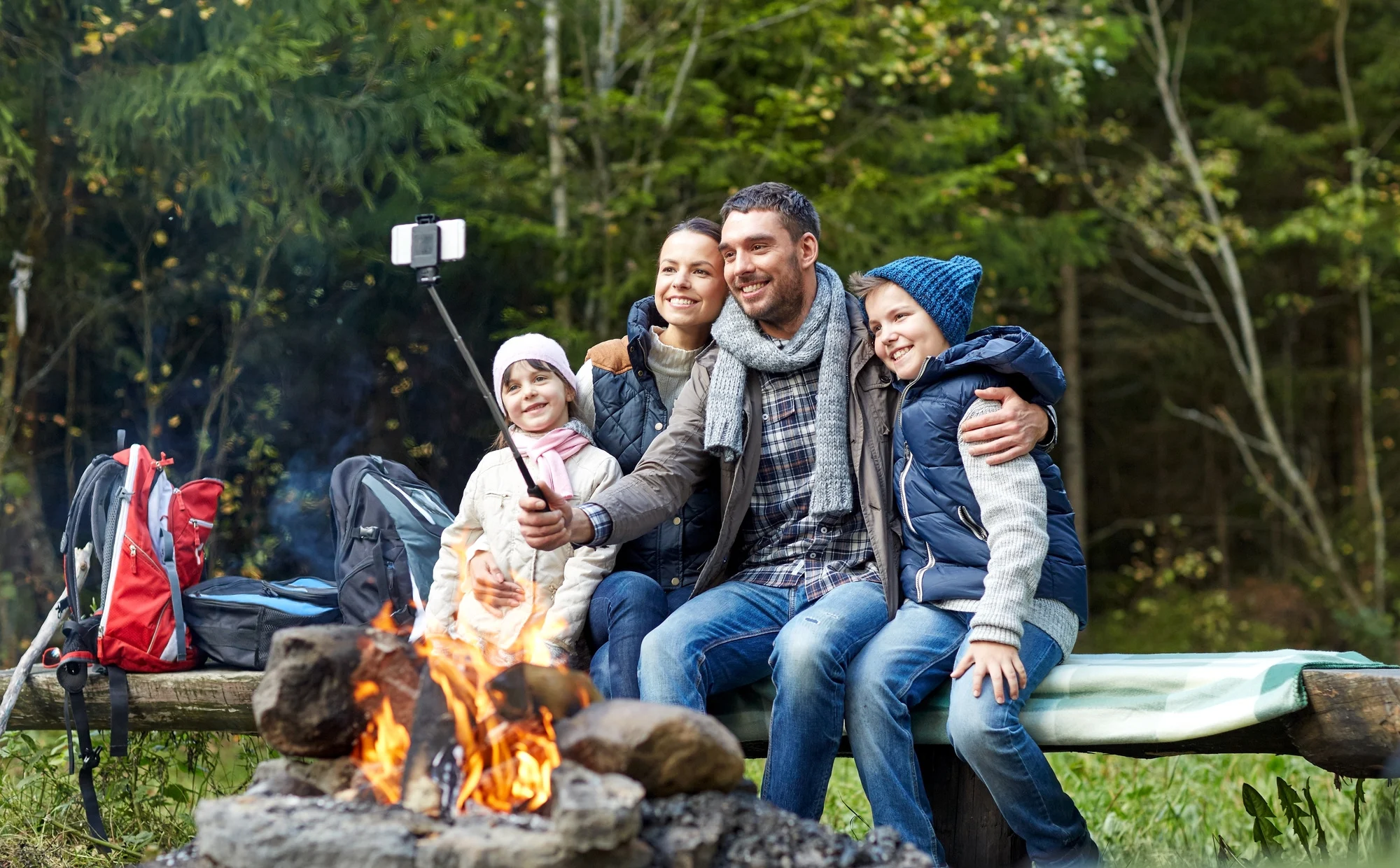 camping, travel, tourism, hike and people concept - happy family sitting on bench and taking picture with smartphone on selfie stick at campfire in woods